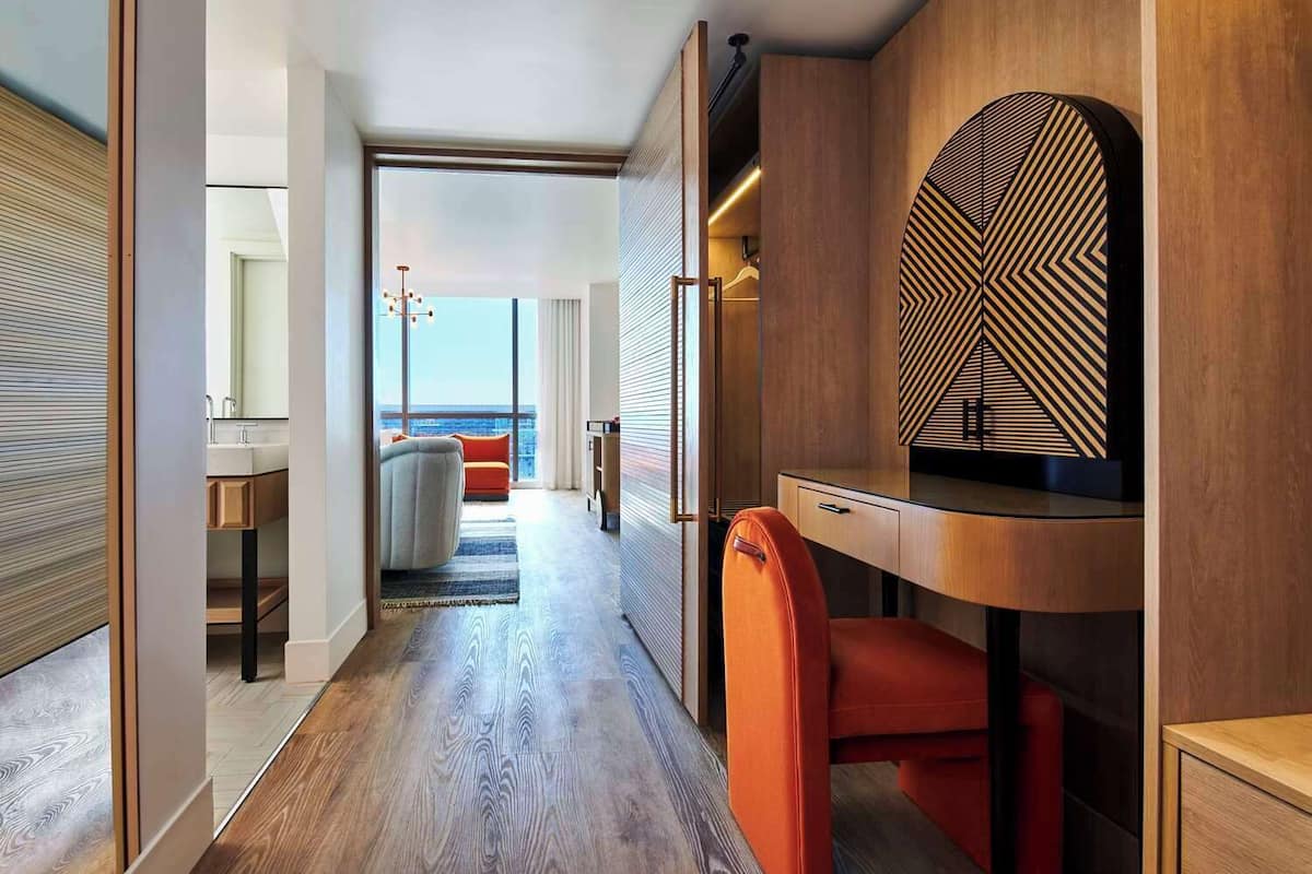 A modern hotel room with a wooden floor, a desk with a chair, a large mirror, and a view of the city through a large window.