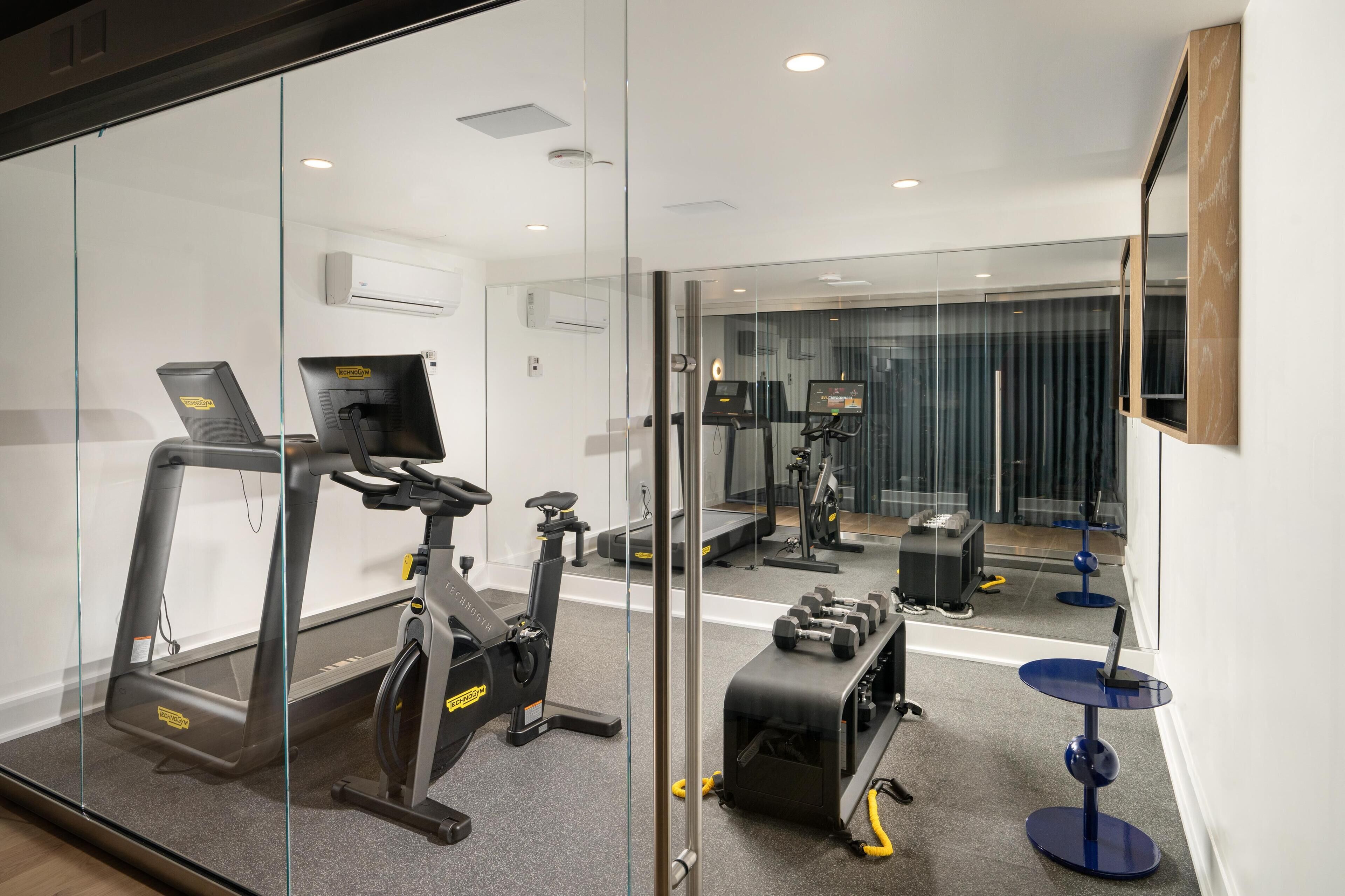 A modern gym with exercise equipment, a mirrored wall, and a small table with a laptop.