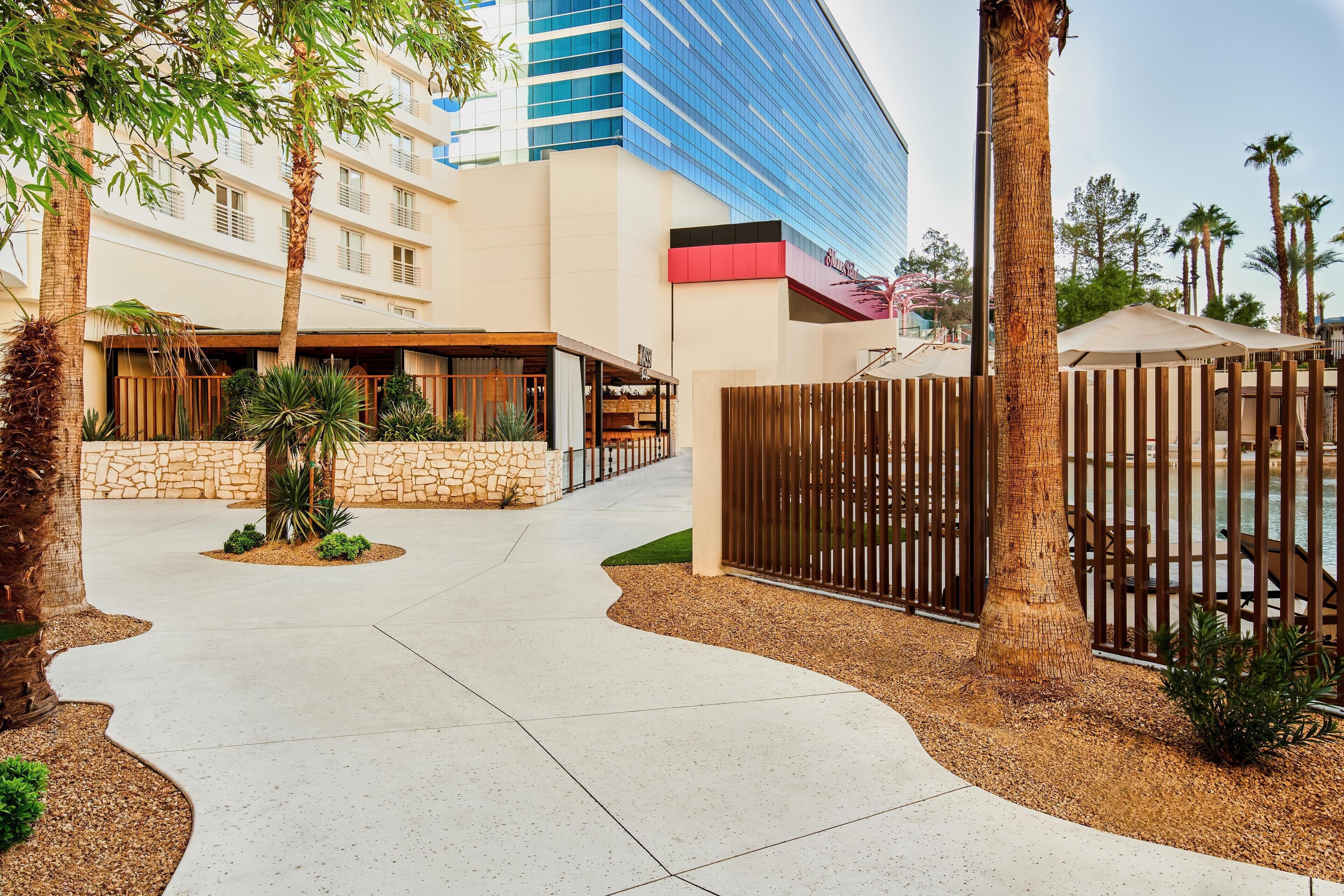 A hotel entrance with a paved walkway, palm trees, and a modern building with a blue glass facade in the background.
