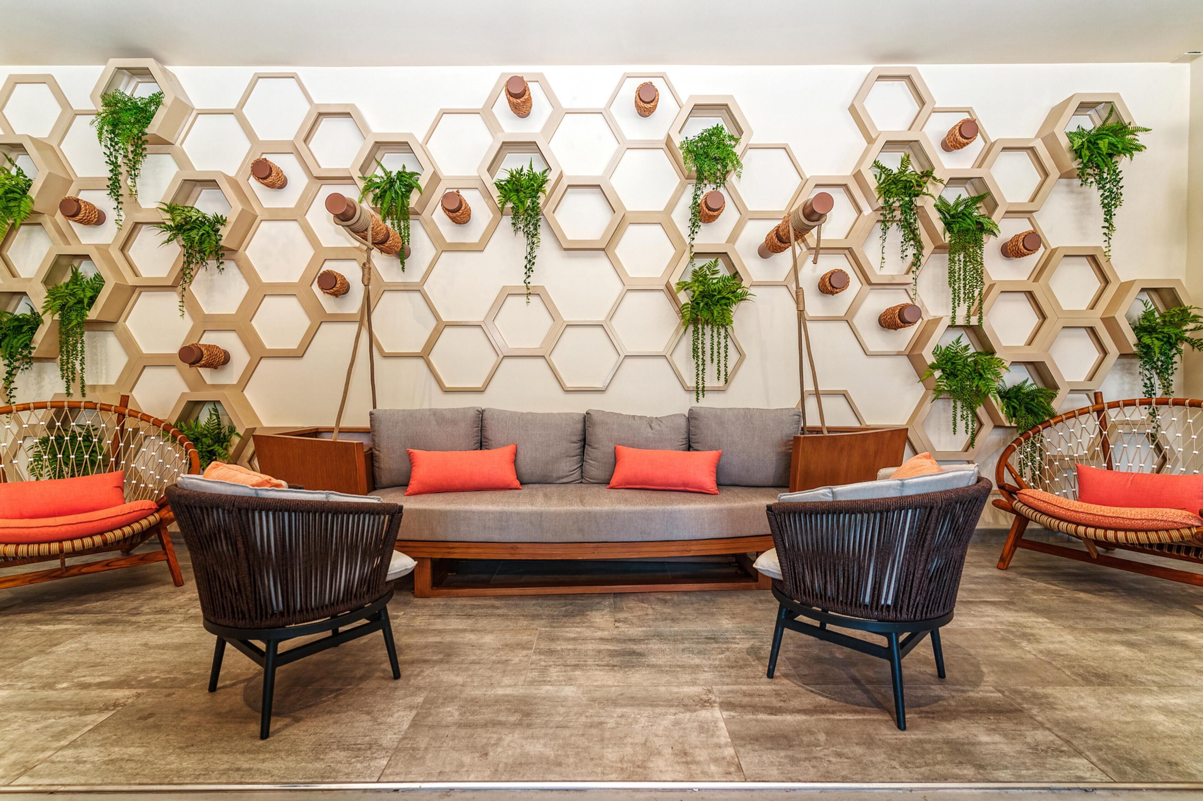 Una sala de estar moderna con un diseño de pared hexagonal, un sofá gris con cojines rojos, dos sillas negras y muebles de mimbre.