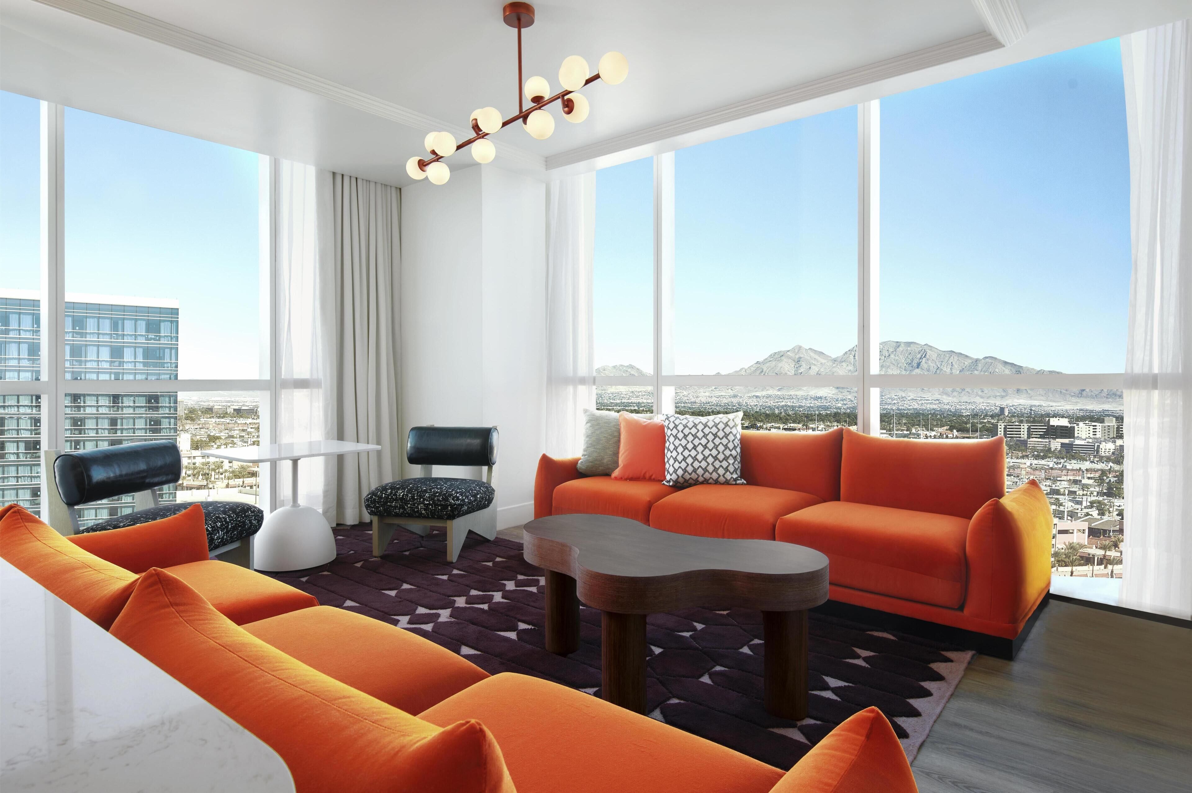 Una sala de estar moderna con un ventanal grande, sofás naranja, una mesa de centro y vistas a un paisaje urbano y montañas.