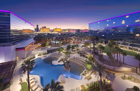 A hotel with a large outdoor pool, palm trees, and a view of the city skyline at dusk.