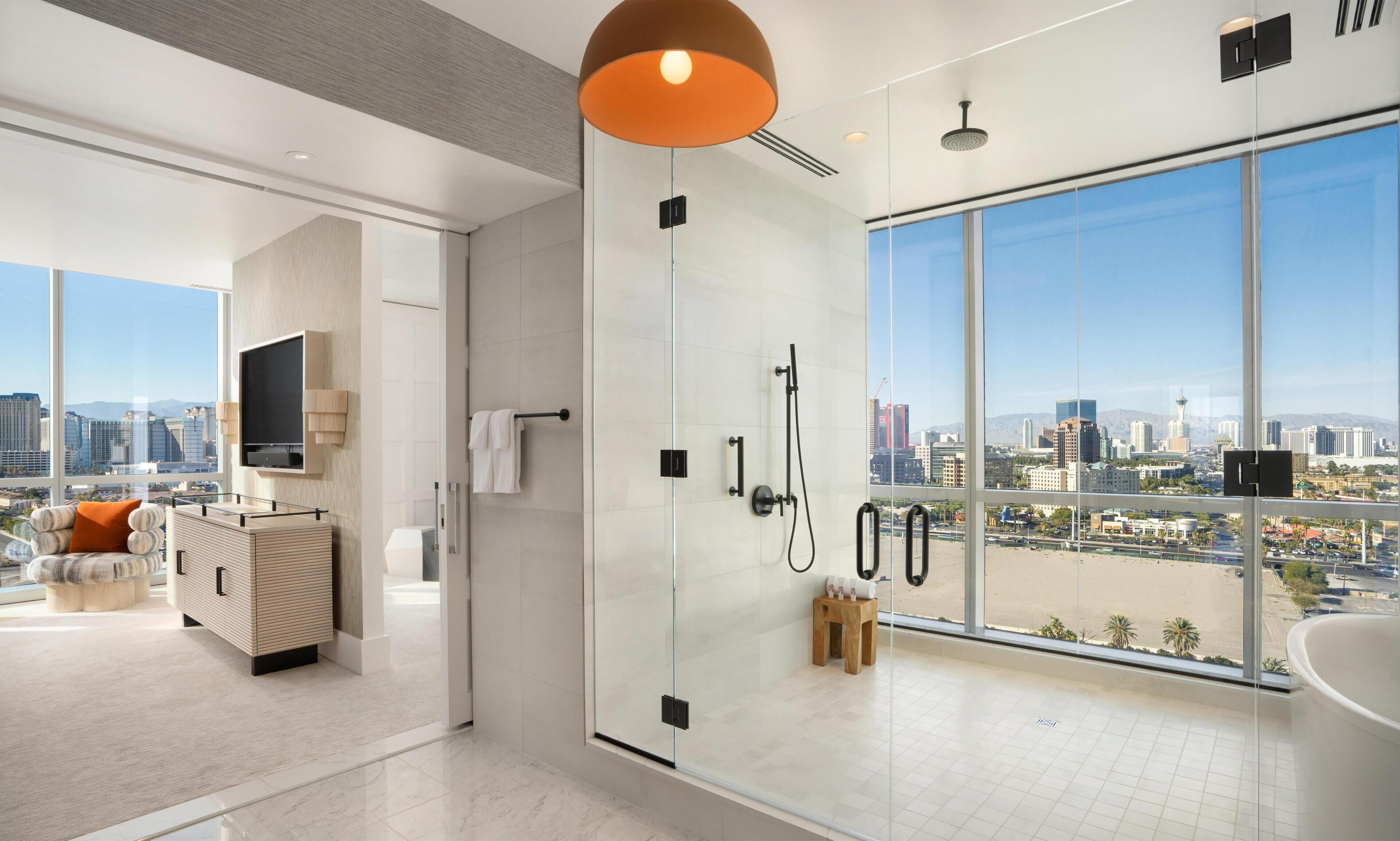A modern bathroom with a glass shower enclosure, a large window offering a city view, and a minimalist design.