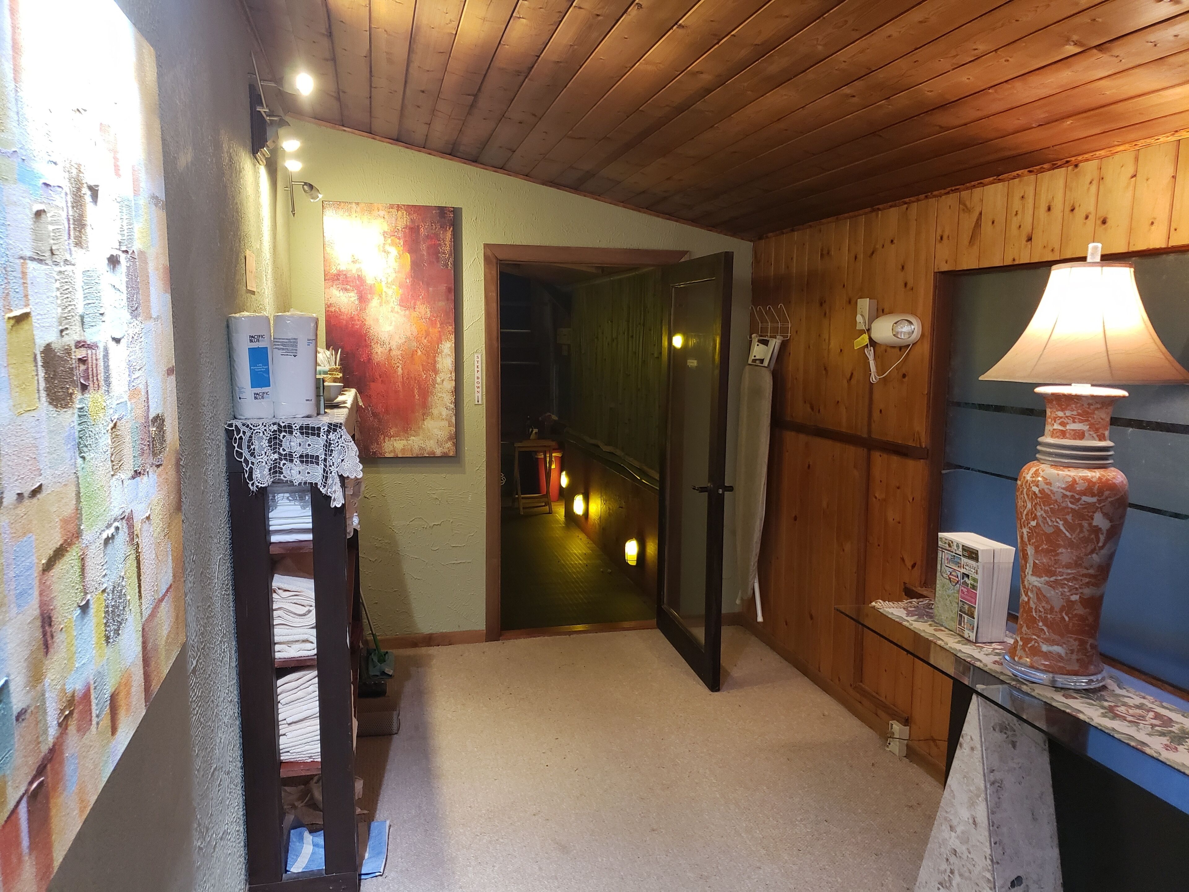 A hallway with a wooden ceiling, a painting on the wall, a lamp, and a door leading to another room.