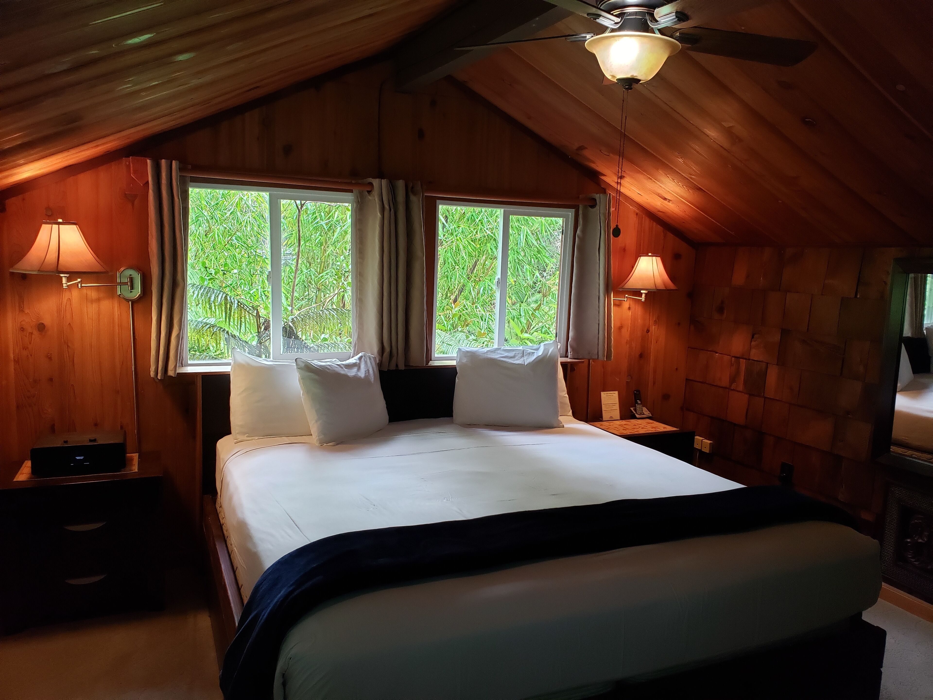 Una camera in legno con un letto, due finestre con tende, una ventola a soffitto e lampade da comodino.