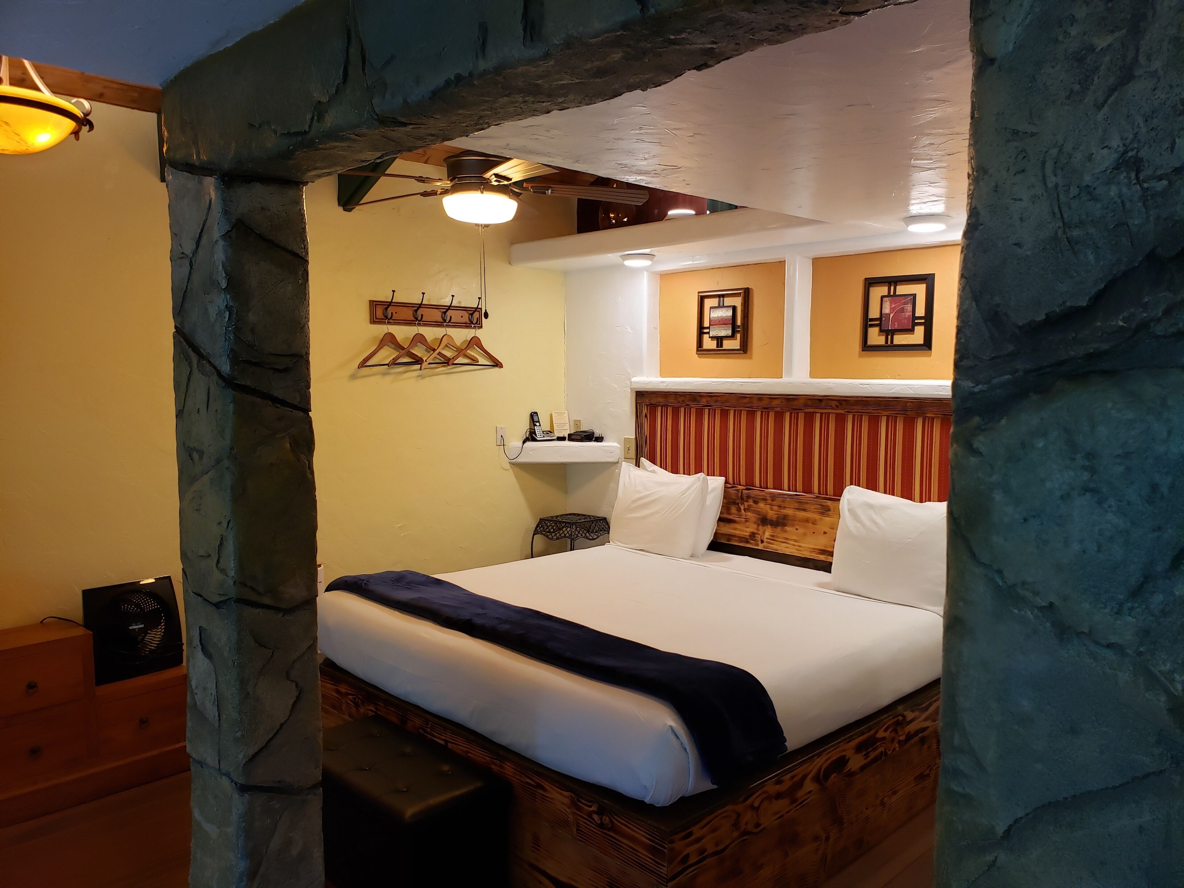 Un dormitorio con un arco de piedra, una cama con ropa de cama blanca, un cabecero de madera, una mesita de noche con una lámpara y un teléfono instalado en la pared.