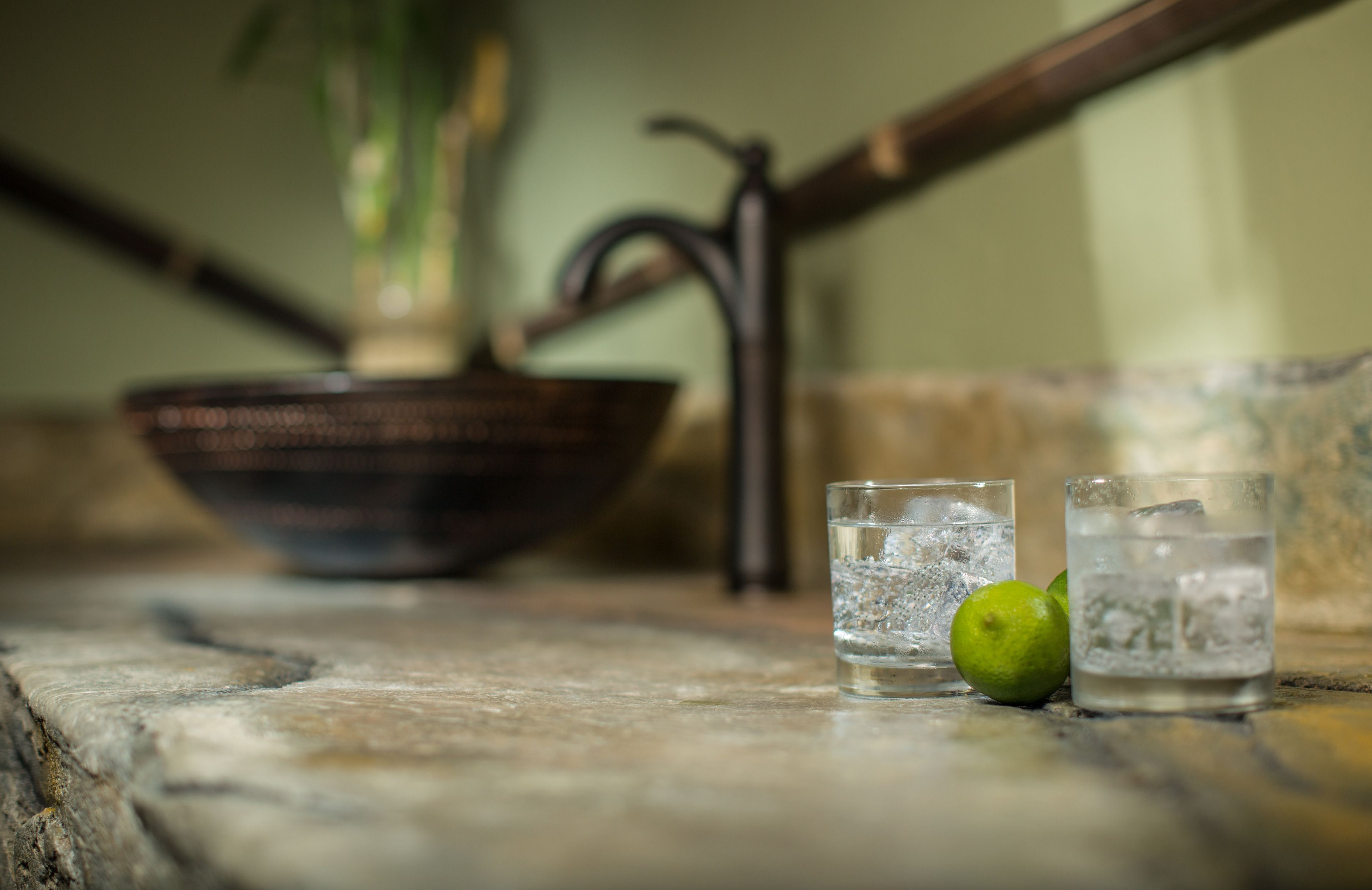 A rustic setting with a metal bowl, two glasses of water, and a lime on a stone surface.