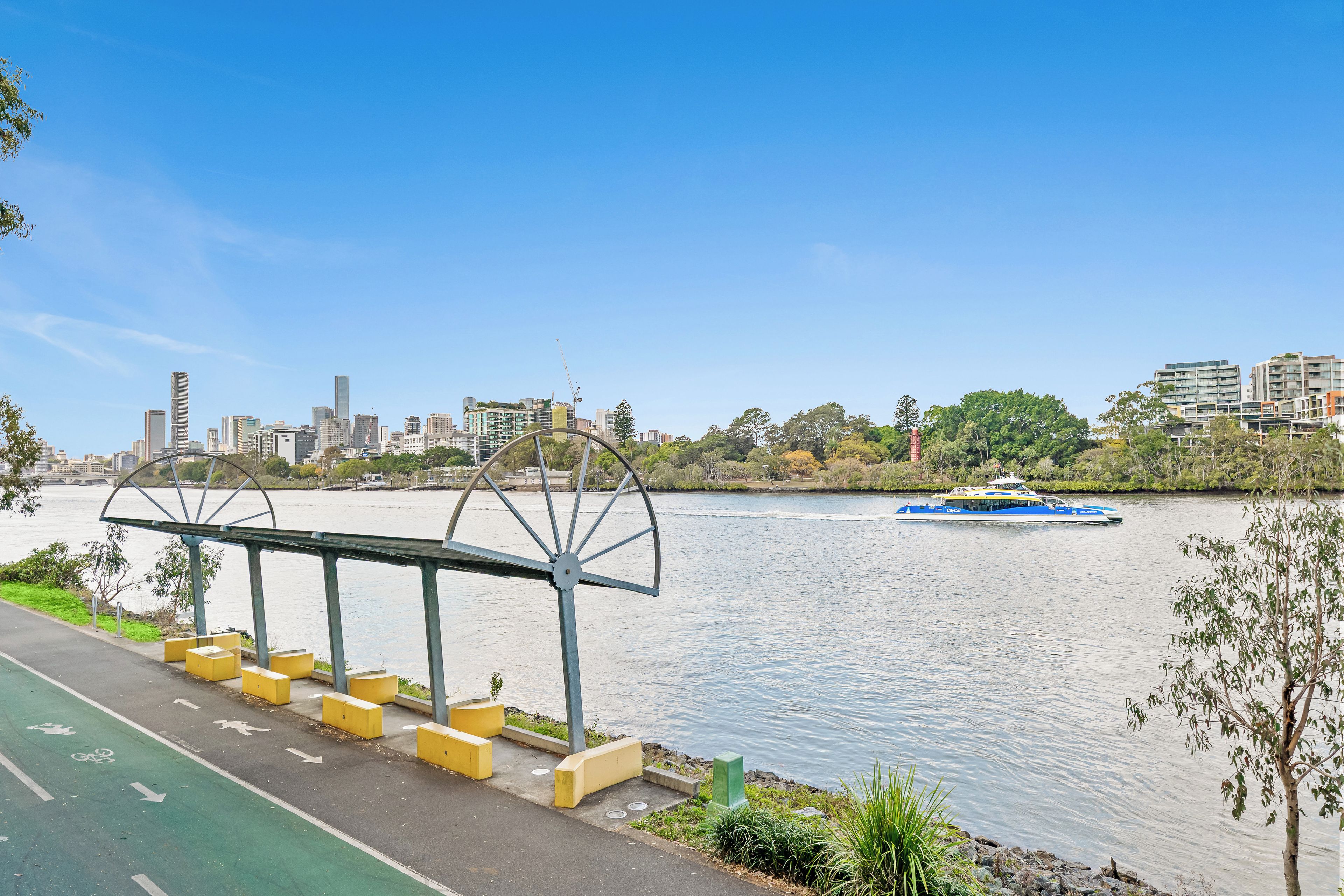 A riverside walkway with a metal shelter and benches, a bike lane, and a city skyline in the background.