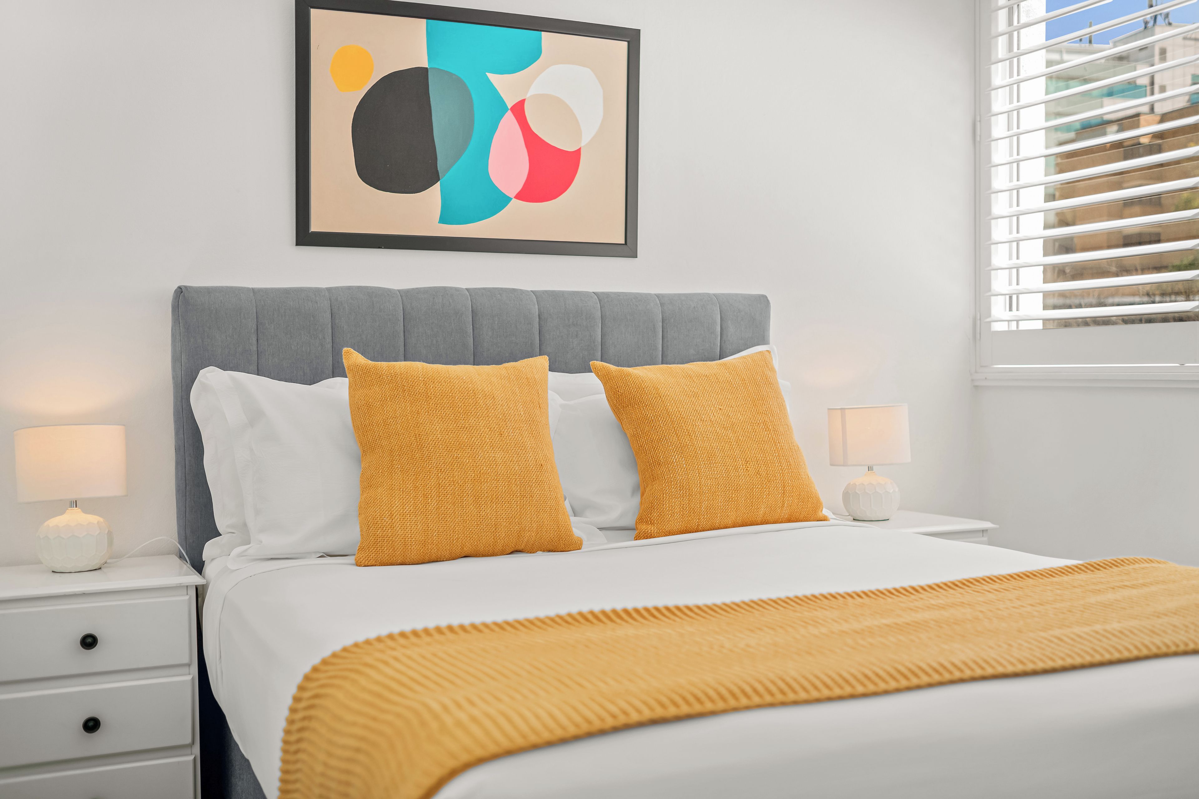Een netjes ingerichte slaapkamer met een bed, twee bedlampen, een nachtkastje en een raam met jaloezieën.