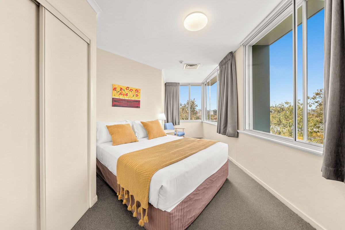 A hotel room with a bed, a painting on the wall, a window with curtains, and a view of the outside.
