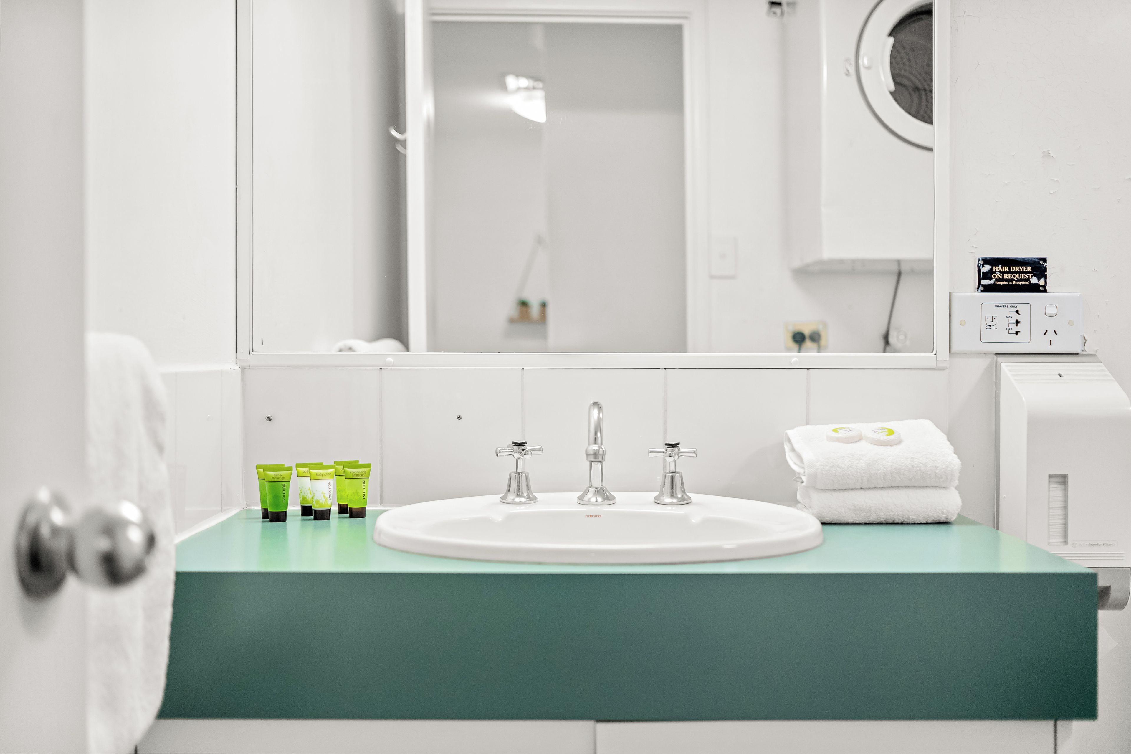 A bathroom with a white sink, two faucets, and a mirror above it. There are green and white toiletries on the counter.