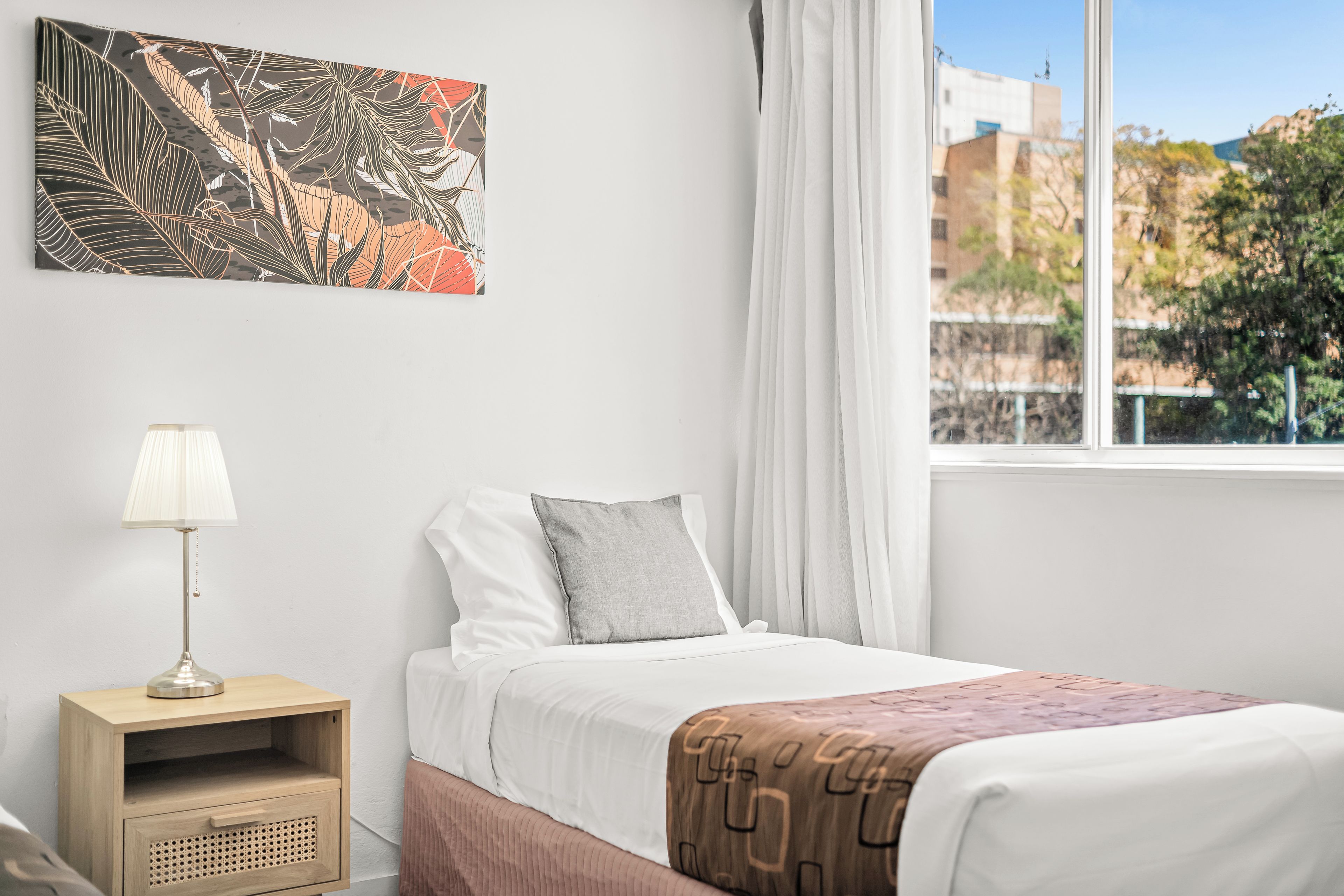 Una camera da letto con un letto, un comodino, una lampada, una finestra con tende e un quadro astratto appeso al muro.
