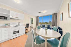 A modern kitchen with white appliances, a dining area with a round table and teal chairs, and a living area with a blue sofa. - Chasely Apartment Hotel (Auchenflower)