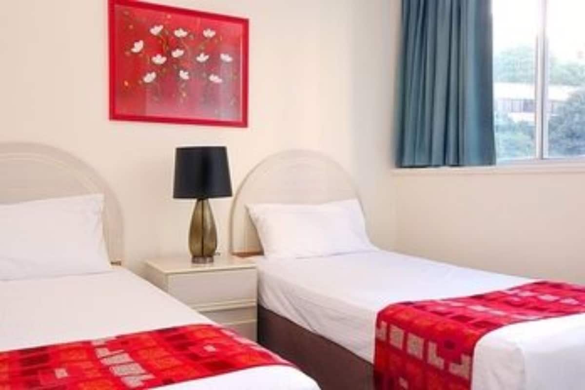 A hotel room with two beds, a nightstand with a lamp, a red artwork on the wall, and a window with curtains.