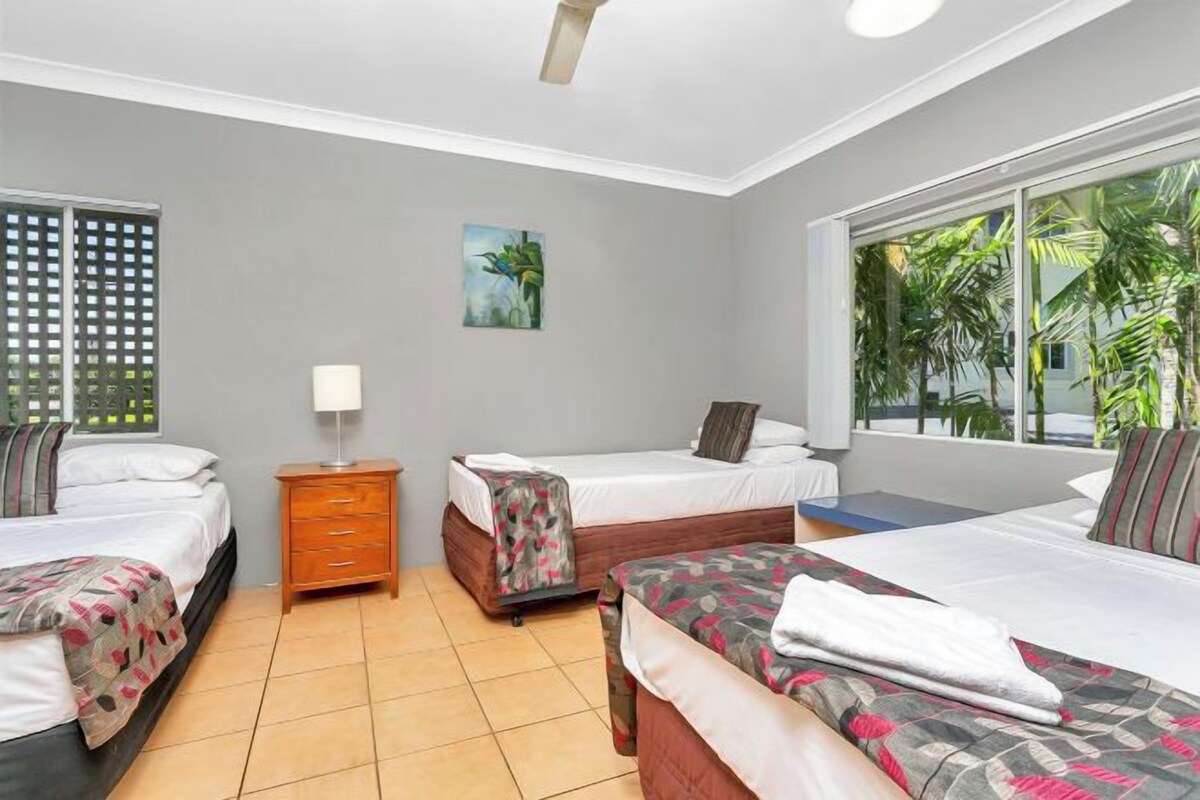 A hotel room with two single beds, a wooden nightstand, a small table with a lamp, and a large window with a view of palm trees.