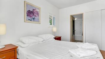 Una cama bien hecha con sábanas blancas, una mesita de noche de madera y una mesita de noche roja, junto a una lámpara fijada en la pared.