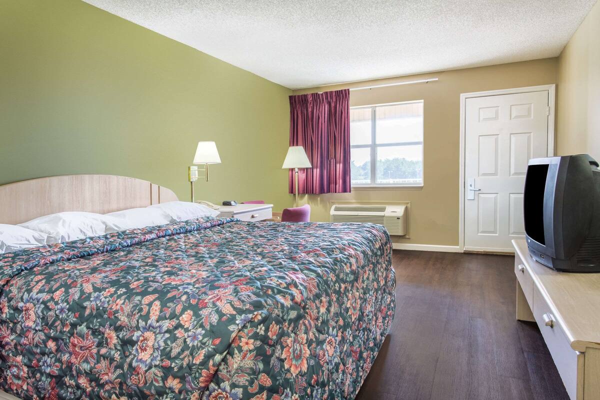 Une chambre d’hôtel avec un grand lit, un téléviseur posé sur une commode, une fenêtre avec des rideaux et une porte.