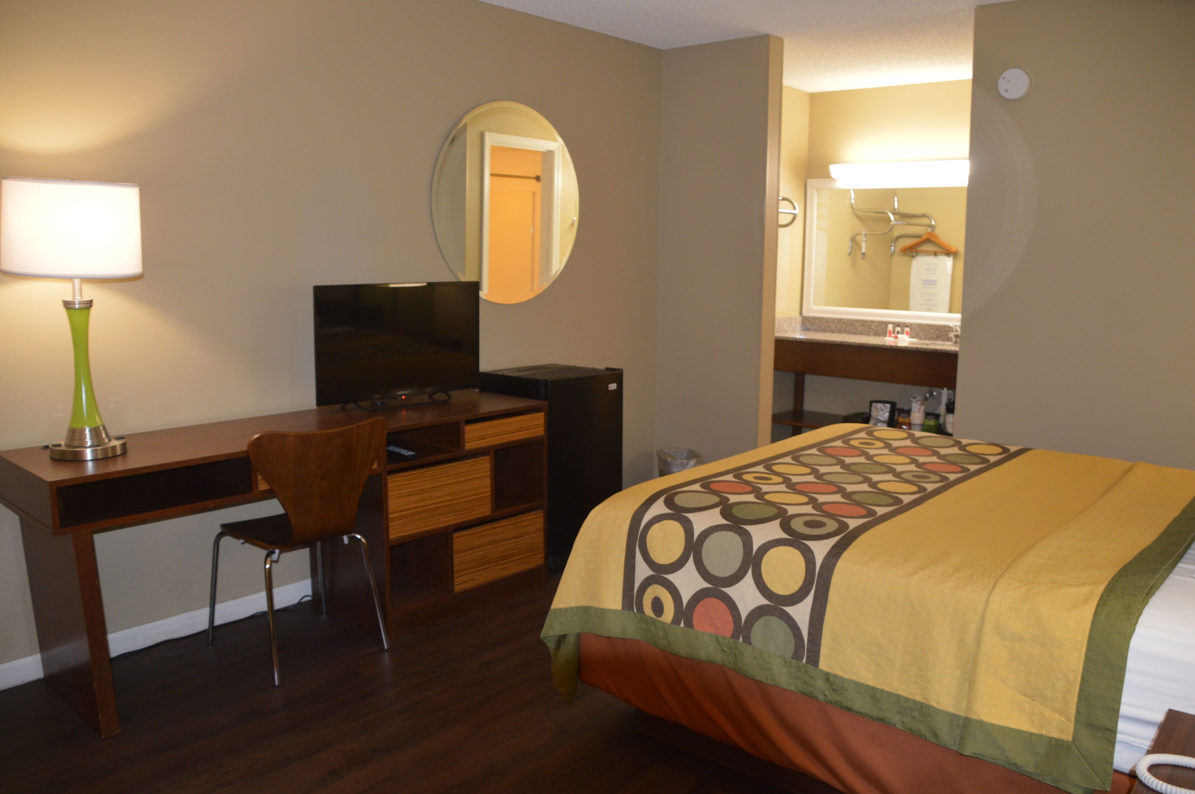 A hotel room with a bed, a desk with a chair, a television, a mirror, and a bathroom with a sink and mirror.