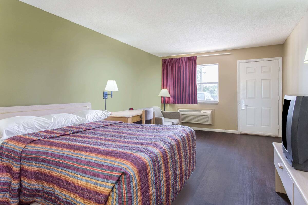 Une chambre d’hôtel avec un lit, un bureau, une chaise, une télévision et une fenêtre avec des rideaux.