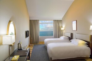 A hotel room with two beds, a TV, a desk with a lamp, a window with a view of the sea, and curtains. - Portemilio Hotel And Resort (Jounieh)