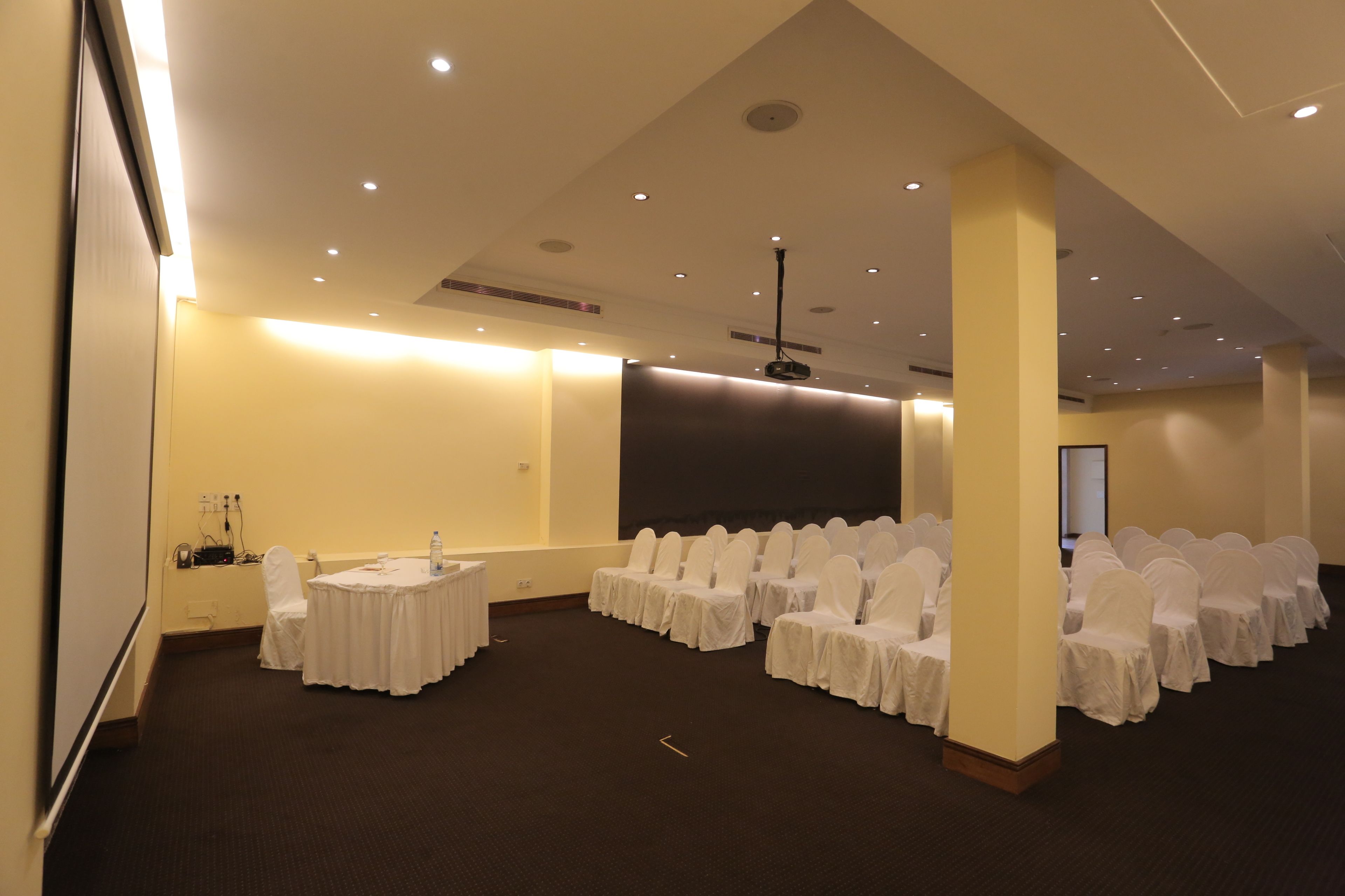 A conference room with rows of white-covered chairs, a projector screen, and a white table.