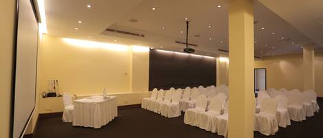 A conference room with rows of white-covered chairs, a projector screen, and a white table.