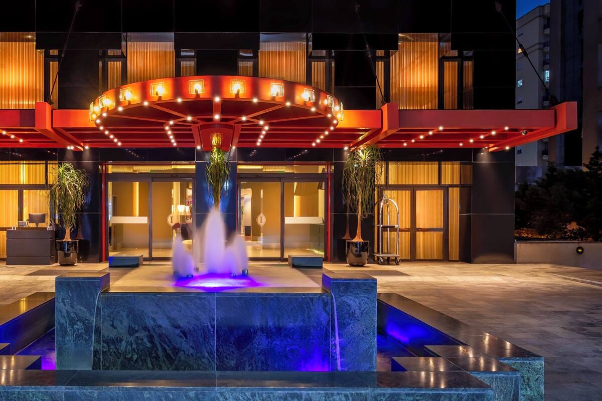 Entrada de un hotel con una fuente, un toldo rojo y una fachada de edificio moderna.