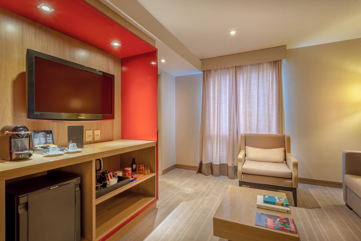A hotel room with a flat-screen TV, a coffee machine, a bookshelf, a seating area with a chair, and a window with curtains.
