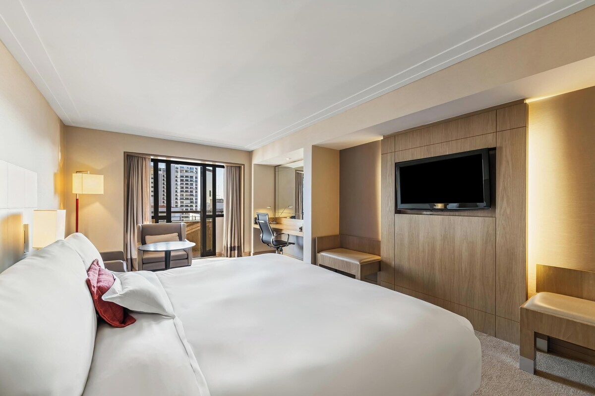 A hotel room with a large bed, a desk, a chair, a TV, and a view of the city.