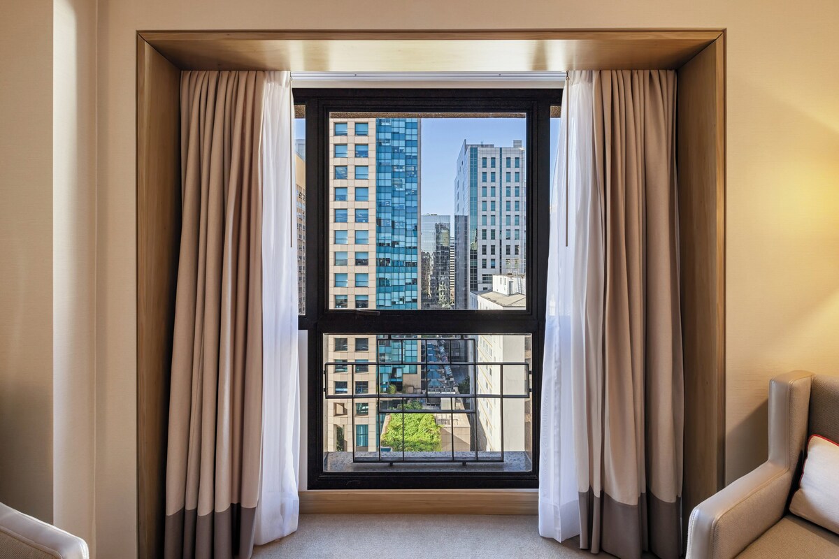 A room with a large window, curtains, a view of tall buildings, and a chair.