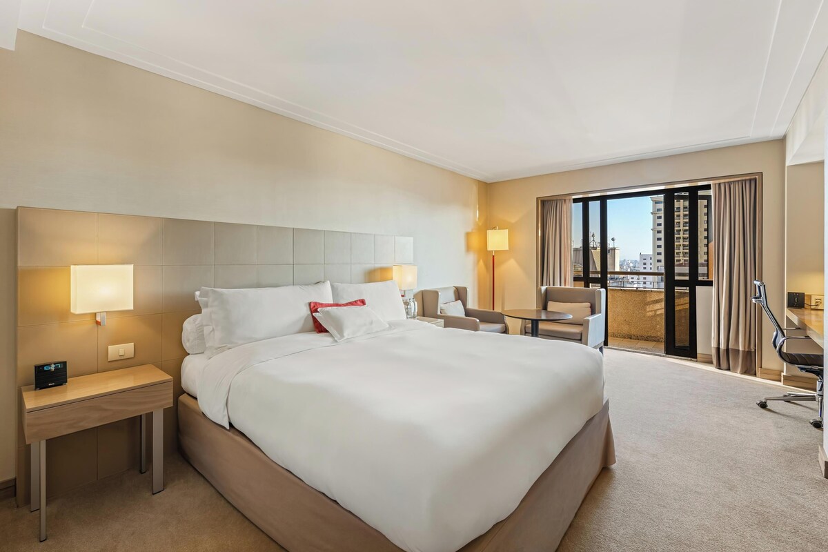A hotel room with a large bed, bedside tables, a desk, a chair, and a view of the city through large windows.