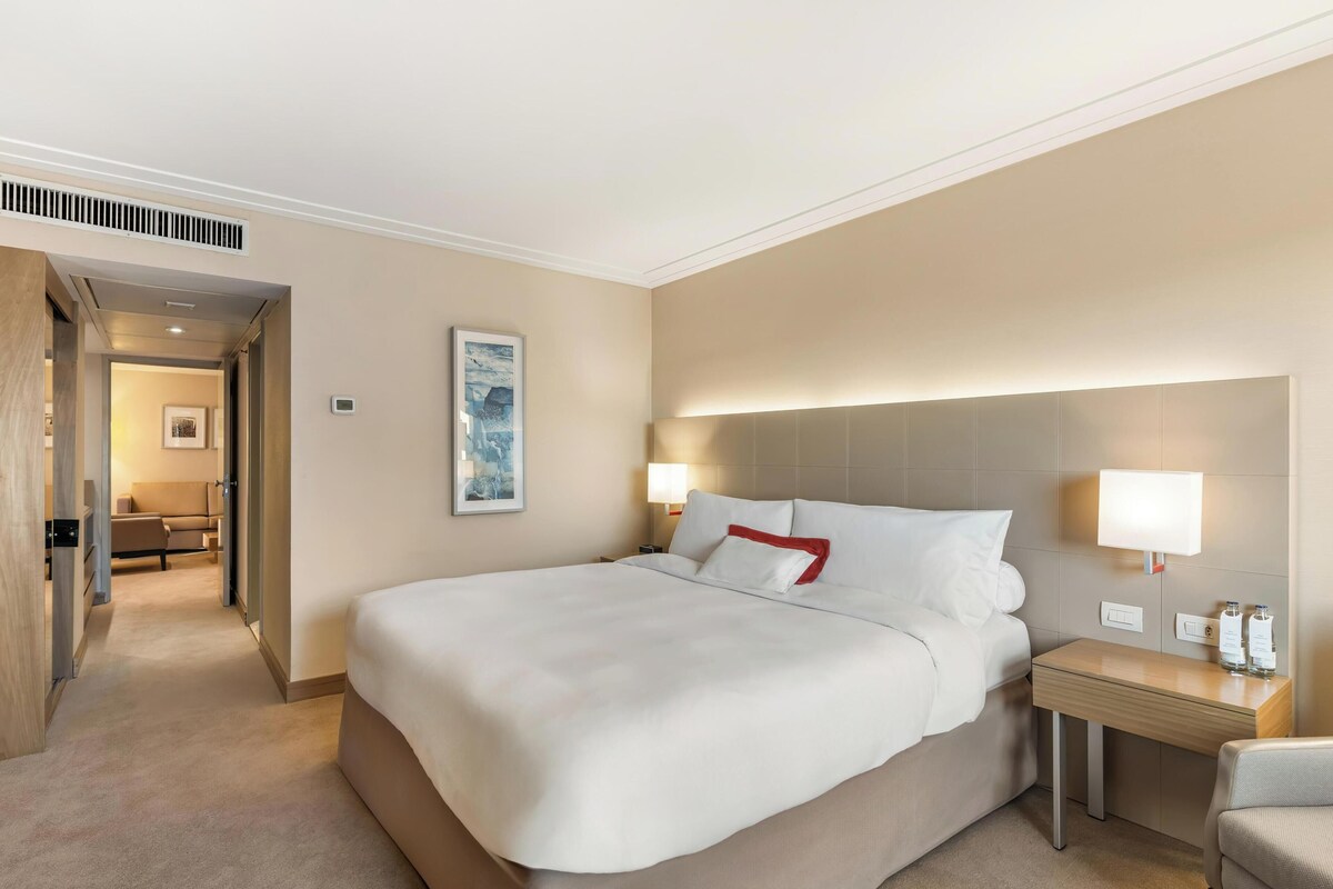 A hotel room with a large bed, bedside lamps, a small table, and a chair. A hallway leads to another room with a sofa.