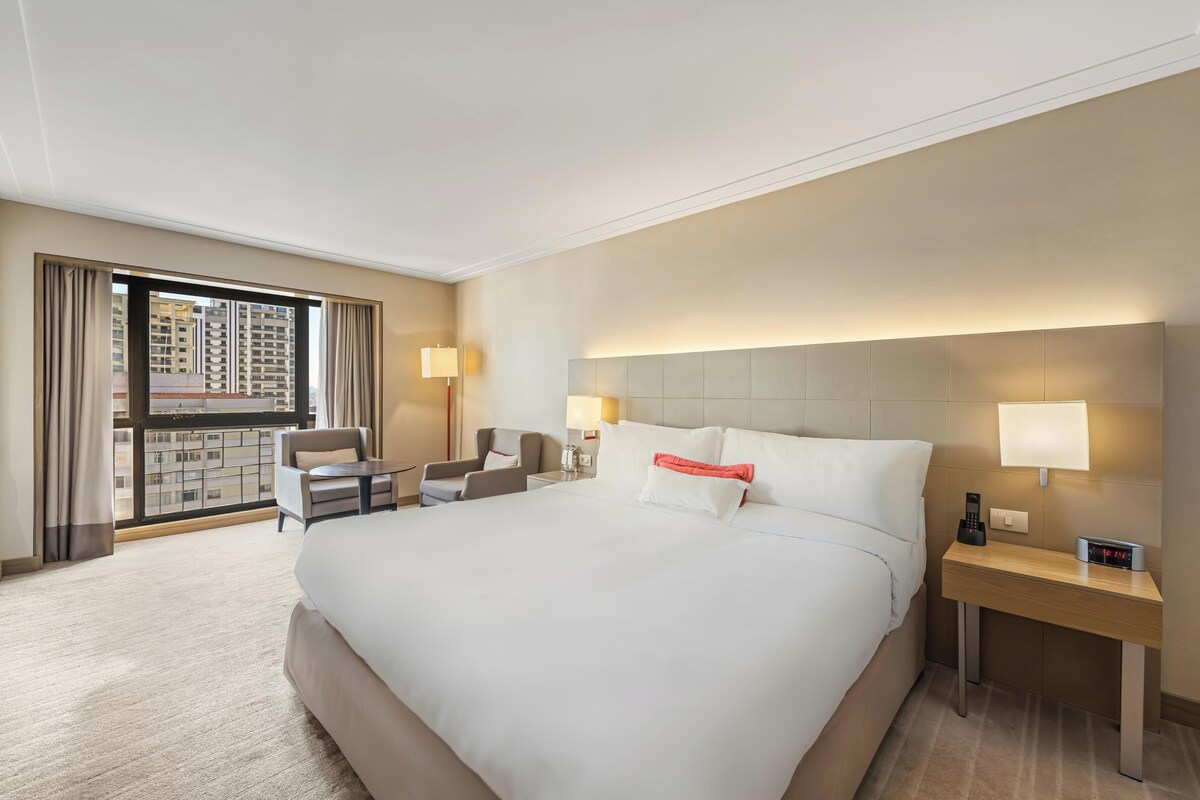 A hotel room with a large bed, two bedside tables, a desk, a chair, and a view of a cityscape through a large window.