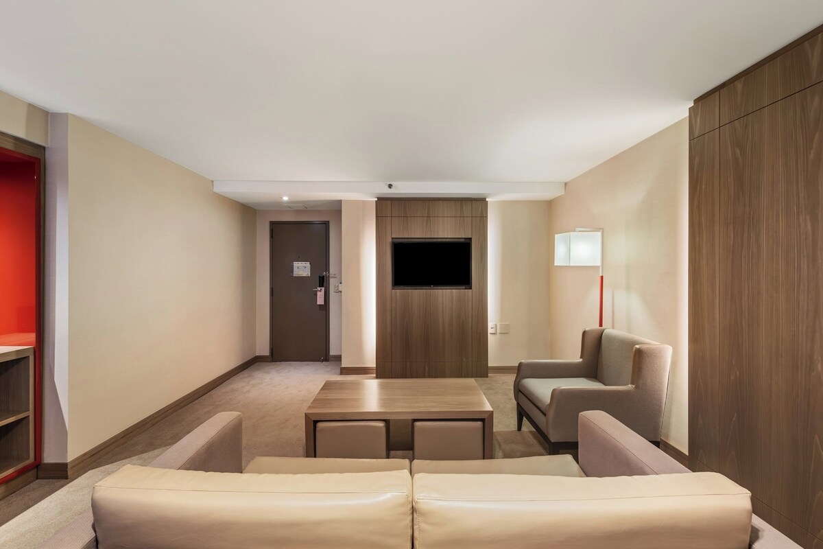 A modern hotel room with a sofa set, a small table, a TV mounted on the wall, and a wooden wardrobe.