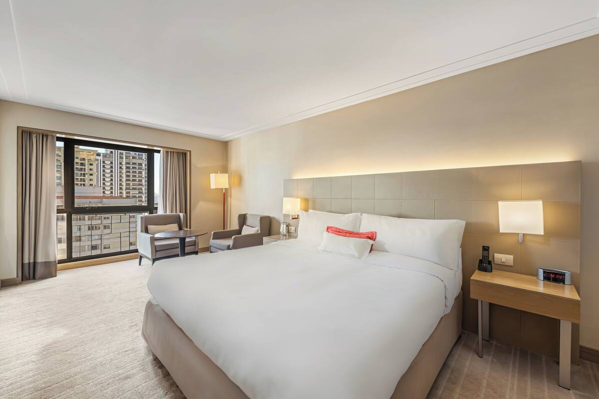 Una habitación de hotel con una cama grande, un escritorio, una silla y vistas de la ciudad desde la ventana.