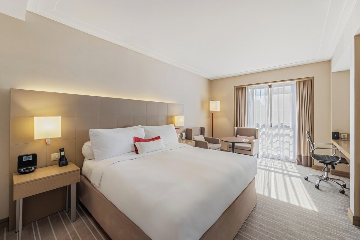 A hotel room with a large bed, bedside tables with lamps, a desk, a chair, and a window with curtains.
