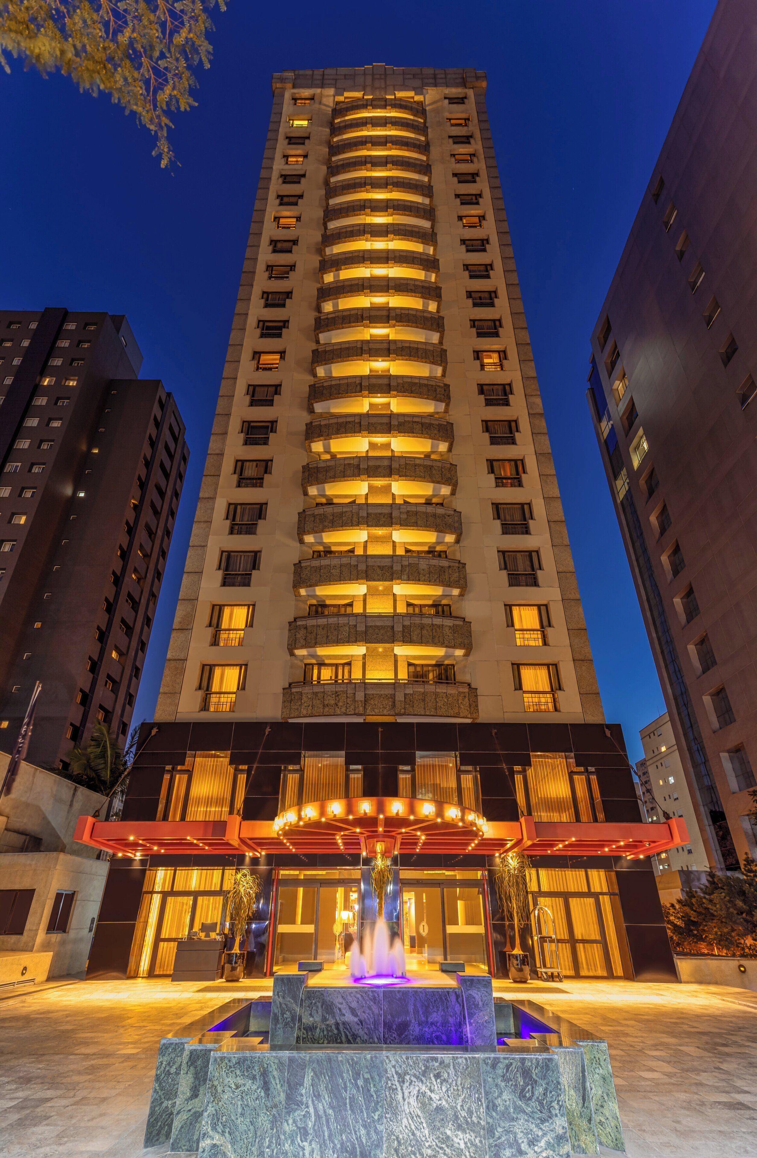 Hotel moderno com vários andares, átrio central e fonte na entrada.