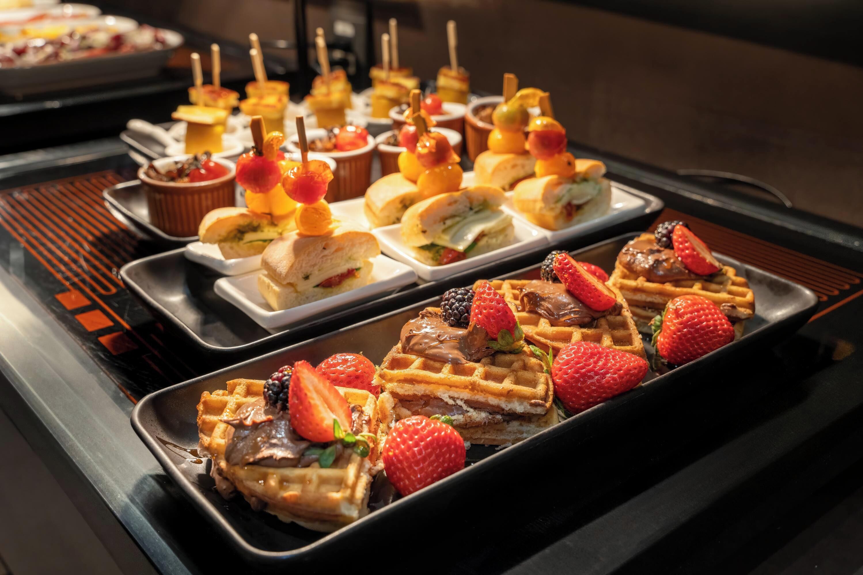 Un buffet con diversi tipi di cibo, tra cui waffle, panini e spiedini di frutta.