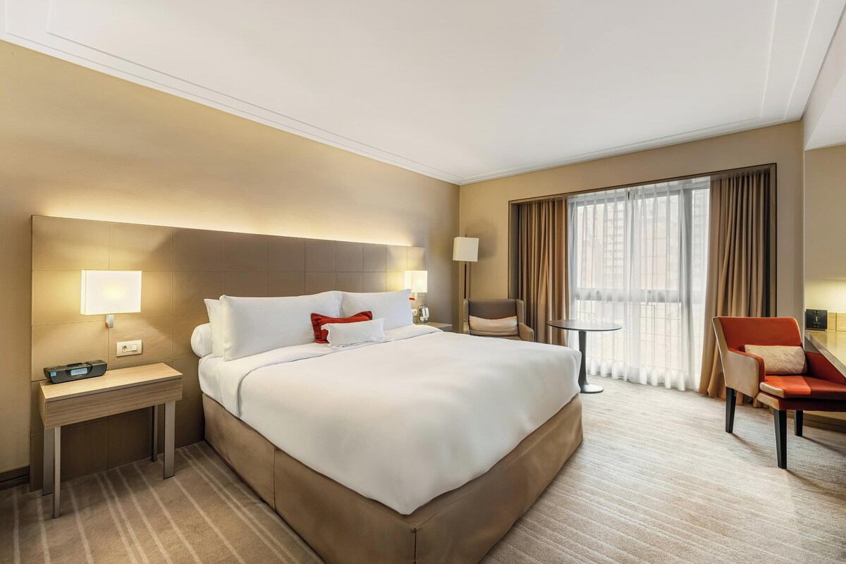 A hotel room with a large bed, bedside tables, a chair, a desk, and a window with curtains.