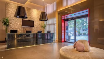 Uma área de recepção moderna com mesa de vidro, duas luminárias pendentes pretas e uma TV grande fixada numa parede texturizada.