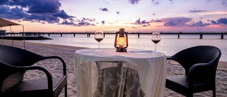 A beachside table with two chairs and a lantern, overlooking a calm sea and a sunset.