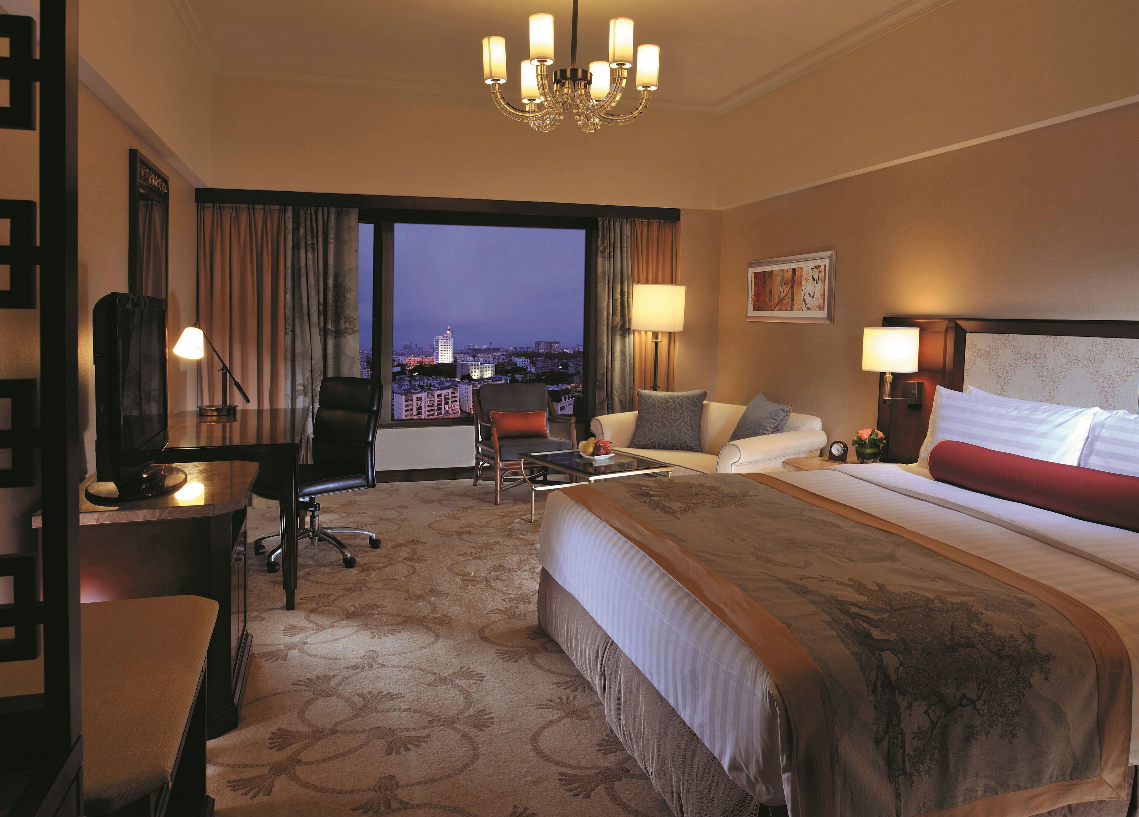 A hotel room with a large bed, two armchairs, a desk with a lamp, a TV, and a view of the cityscape at night.