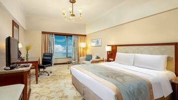 A hotel room with a large bed, a desk with a computer, a chair, a TV, and a window with a view of the sea.