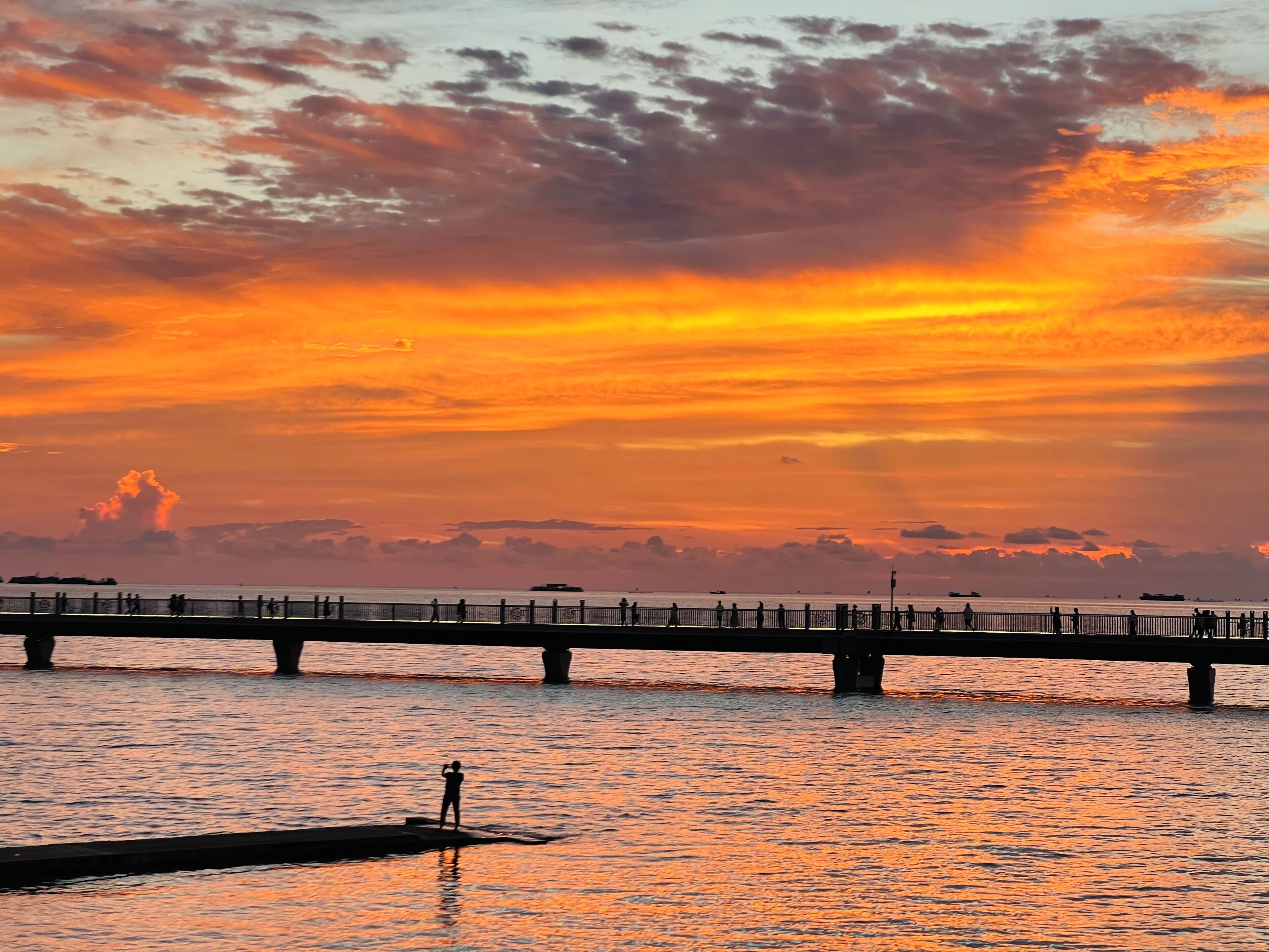 Un coucher de soleil sur une étendue d’eau, avec un pont et des personnes qui se promènent.