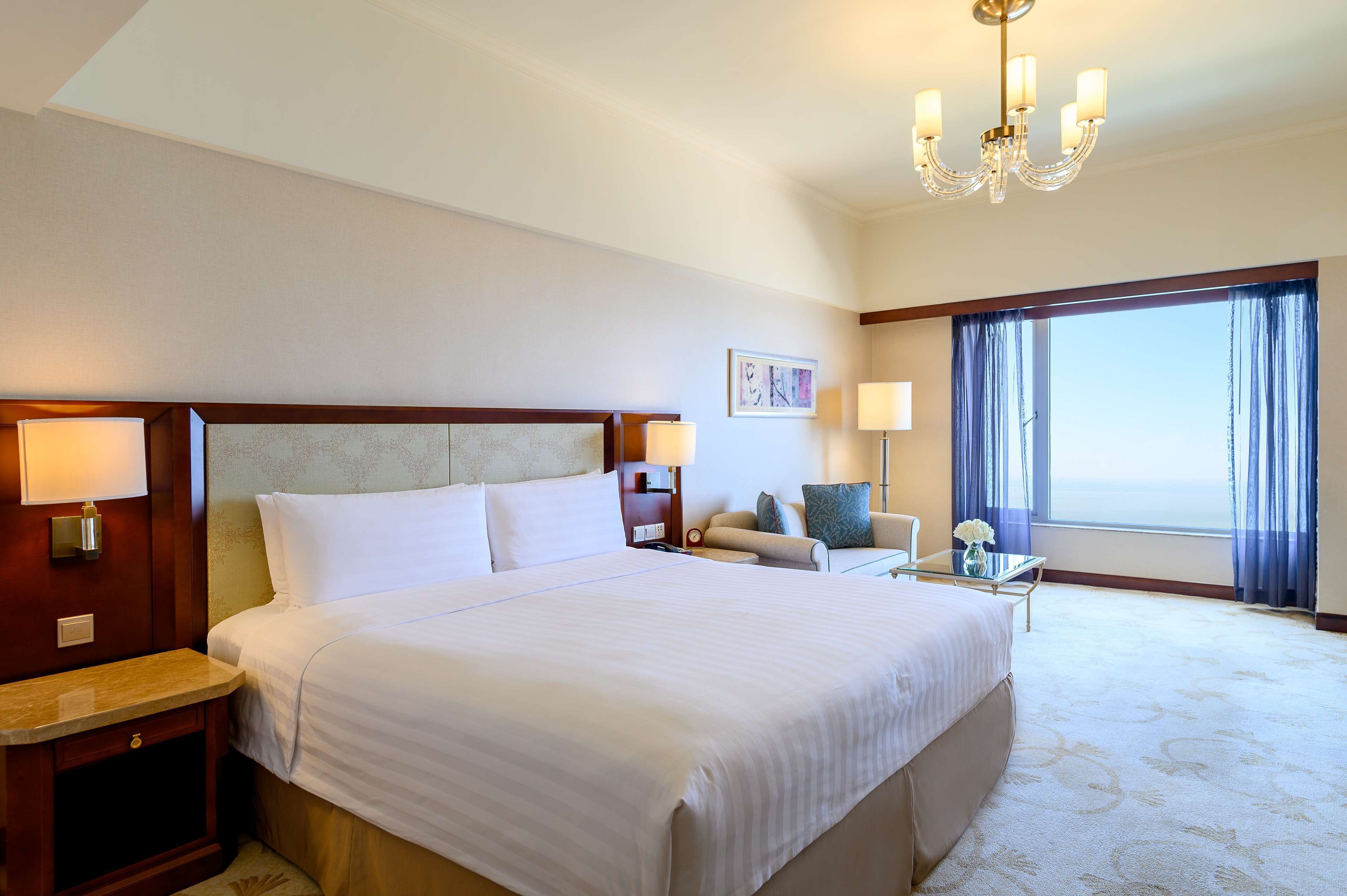 Habitación de hotel con una cama grande, un escritorio con lámpara, una silla y vista al océano a través de la ventana.
