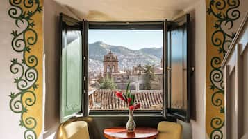 A room with a bed, a dining table with chairs, a vase with flowers, and a view of a cityscape through an open window.