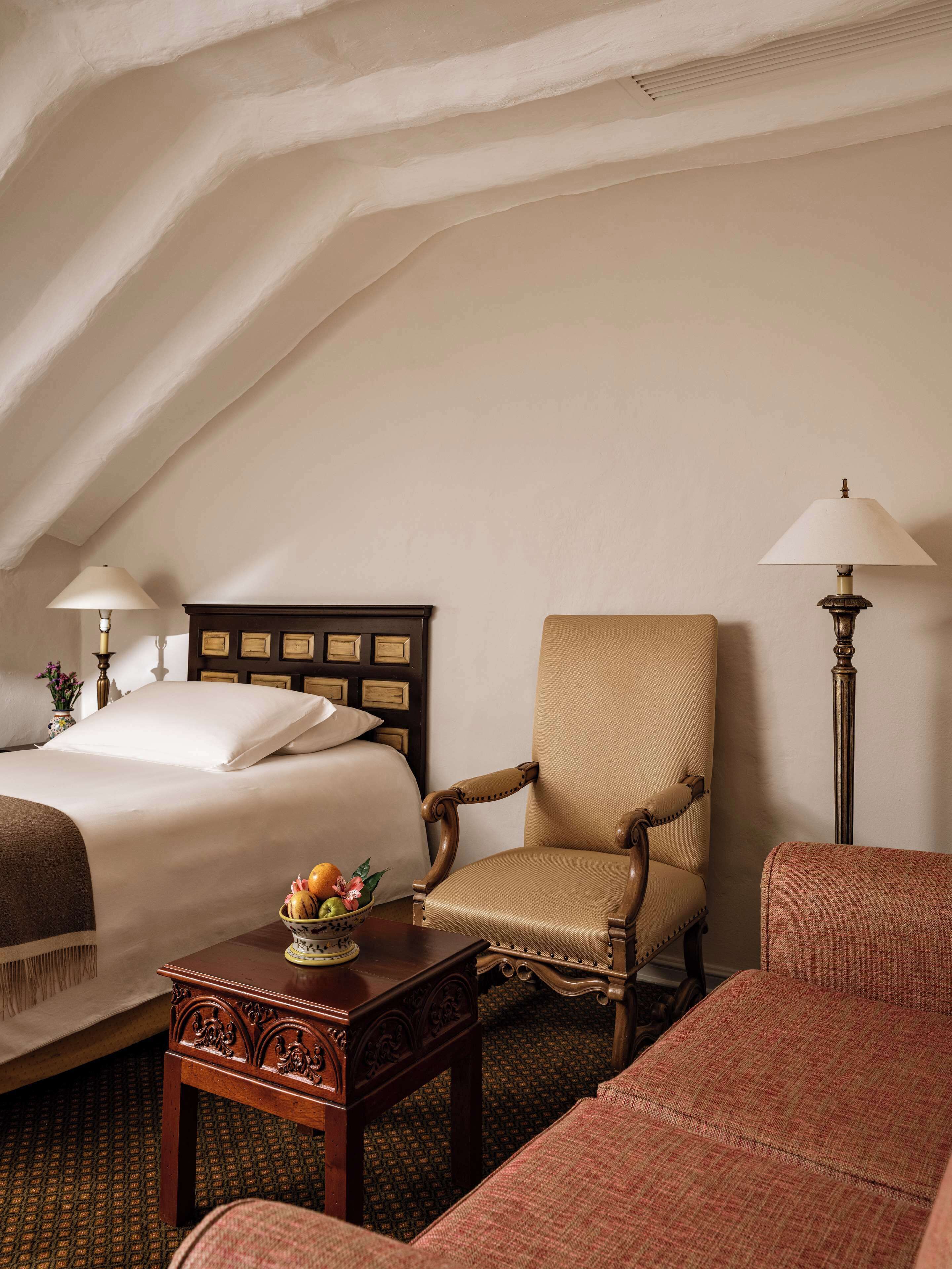 Une chambre d’hôtel avec un lit, deux chaises, une petite table sur laquelle se trouve un panier de fruits, et deux lampes.