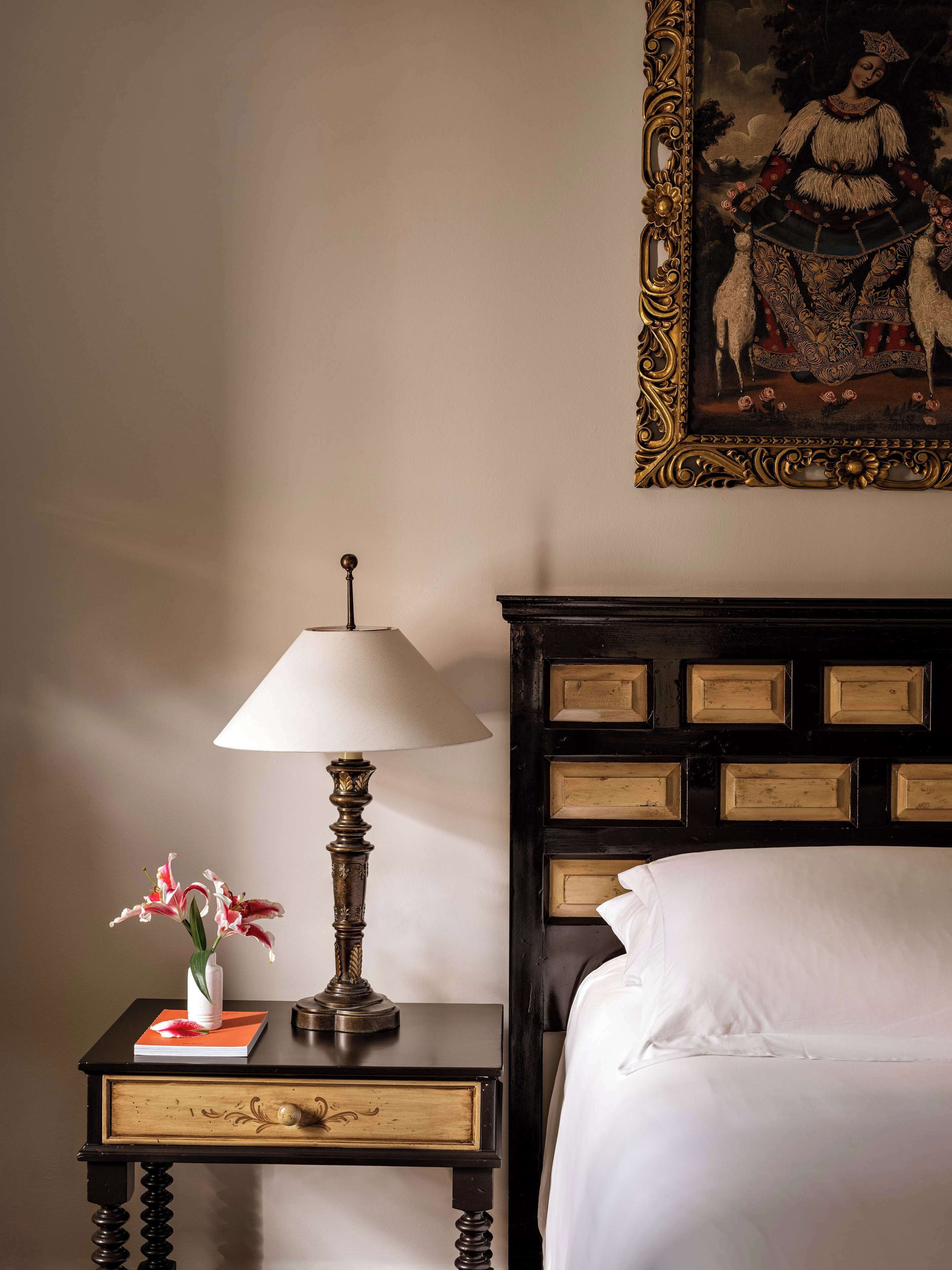 Una cama bien tendida con sábanas blancas, una mesita de noche de madera oscura con una lámpara, un cuadro enmarco y un jarrón con flores.