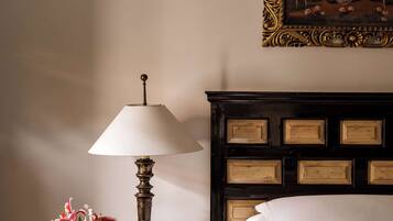 Una cama bien tendida con sábanas blancas, una mesita de noche de madera oscura con una lámpara, un cuadro enmarco y un jarrón con flores.