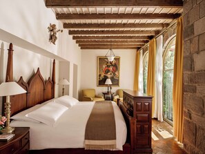 Un dormitorio con una cama grande, cabecera de madera, dos lámparas de noche, un cuadro en la pared, una silla, un escritorio y una ventana con cortinas.